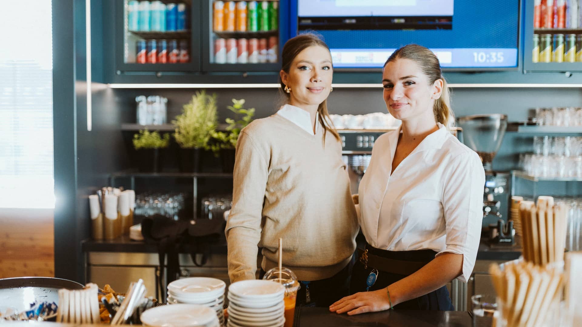 Two staff members smile behind a stylish coffee bar, surrounded by cups, drinks, and fresh herbs – a welcoming moment from the event area.